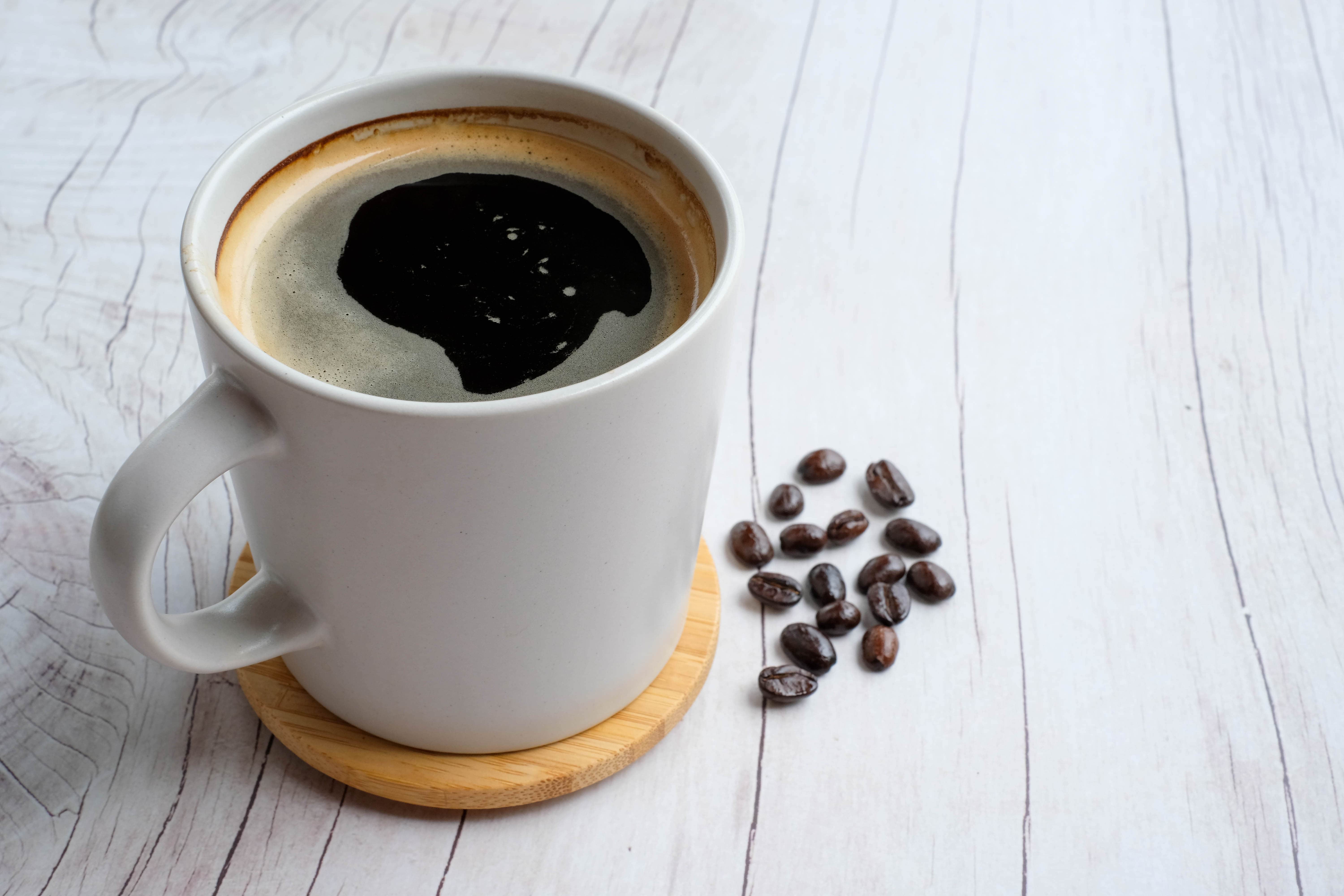 americano black coffee in a white mug on a wooden coaster with loose coffee beans on the side