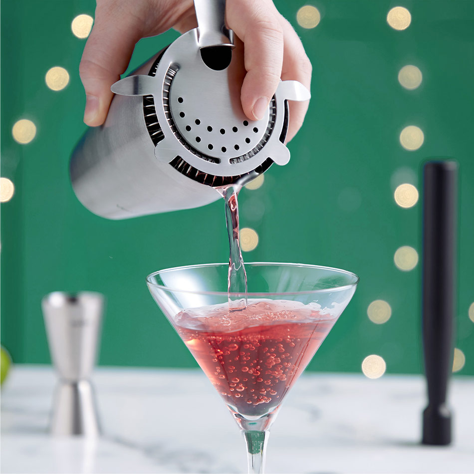 hand pouring a red cocktail into a martini glass with a stainless steel cocktail shaker